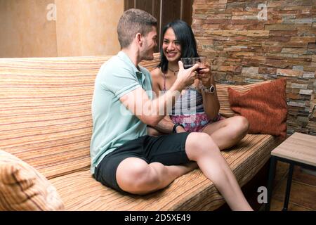 Smiling couple making a toast with wine to celebrate her love. Escapade celebration relaxing at the sofa. Stock Photo