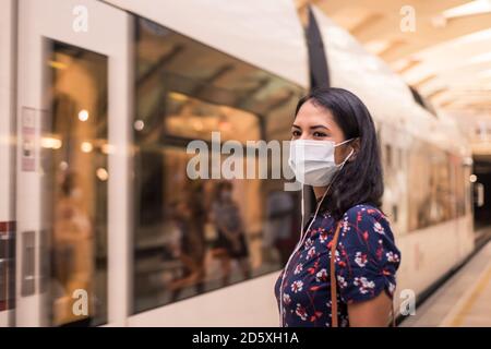 Young latin woman wears face mask and listening to music with earphones waiting for the train in the station. Stock Photo