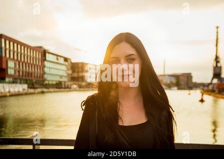 Germany, Muenster, portrait of young woman standing in front of city harbour at backlight Stock Photo