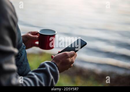Man standing at lake, drinking coffee, using smartphone Stock Photo
