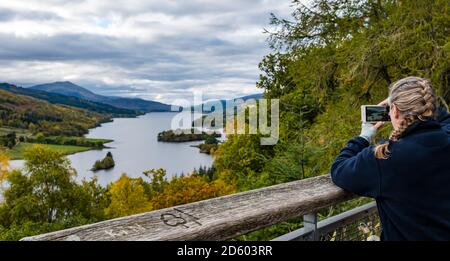 Perthshire, Scotland, United Kingdom, 14th October 2020. UK Weather: Autumn colours. The trees across Perthshire display stunning gold and orange colours on a day that alternated between rain and sunny intervals. Pictured: Queen's View, Loch Tummel with Schiehallion mountain peak in the distance as a woman takes a photo with a mobile phone at the viewpoint