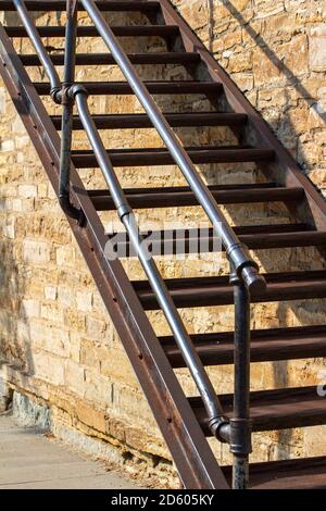 Sturdy metal fire escape staircase along an old white rough textured stone wall background, in full sunlight with copy space Stock Photo