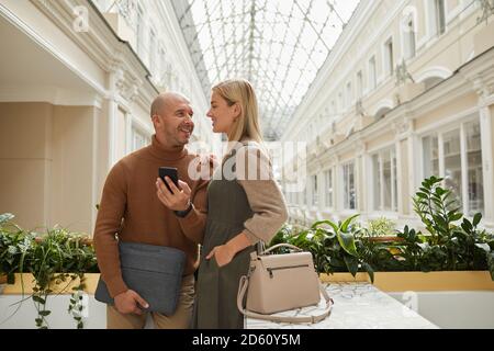 Two friends talking to each other and using mobile phone while standing at the restaurant Stock Photo