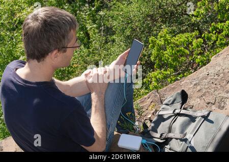Man on a hike uses smartphone while charging from the power bank sitting on the rock over the forest. Stock Photo