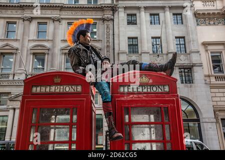 Punk rocker with mohican hair sits relaxing on top of red telephone boxes in central London, England, United Kingdom Stock Photo