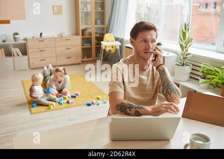 Father sitting at the table and talking on the phone while his family playing in the background in the room Stock Photo