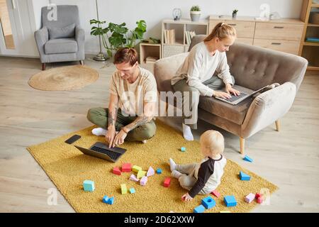 Two parents using laptop computers and working online at home while their son playing with toys on the floor Stock Photo