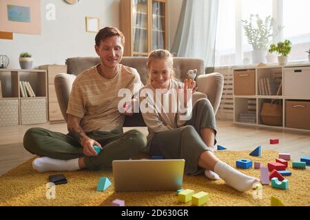 Young couple sitting on the floor smiling and waving looking at monitor of laptop they talking online at home Stock Photo