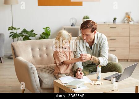 Father helping to his little son doing homework they sitting on sofa at the table in the room Stock Photo