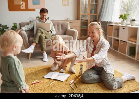 Happy mother playing on the floor together with her children while father working on laptop sitting on sofa at home Stock Photo