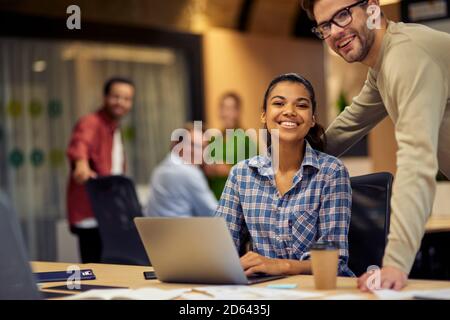 Two young multiracial colleagues looking at camera and smiling while working together on laptop in the modern office or coworking space. Teamwork, cooperation, business concept Stock Photo