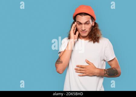 Freestyle. Curly-haired guy wearing hat standing isolated on blue touching stomach and head concerned Stock Photo