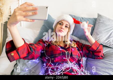 Excited young woman in santa hat wrapped in a garland making selfie photo on mobile phone lying on the bed Stock Photo