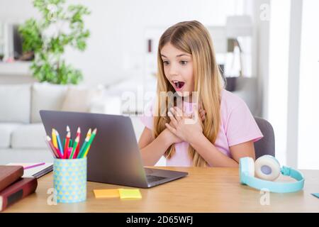 Photo of astonished kid girl study remote use laptop impressed homeschool teacher news information touch hands chest sit comfort table desk in house Stock Photo