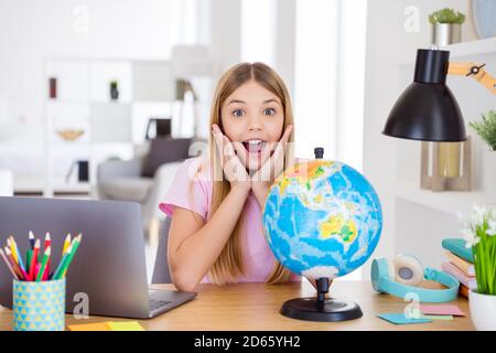 Photo of astonished kid girl study geography remote impressed new continent information scream touch hands face sit comfort table desk in house Stock Photo