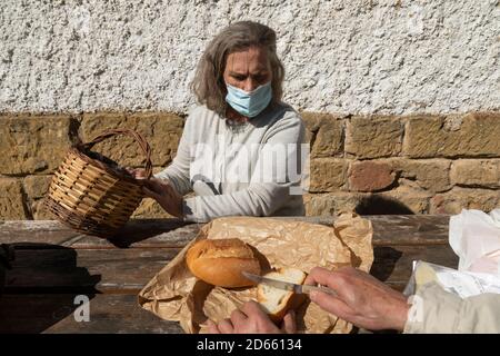 A senior adult woman, wearing a face mask, chooses the amount of bread she wants, before eating at the mountain hut of the fountain of Artica, in Lues Stock Photo