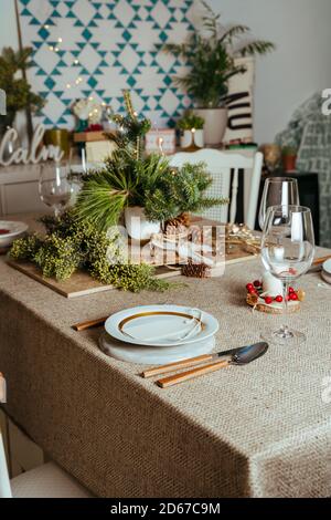 Homely christmas table setting, decorated with pine branches and rustic tablecloth in the living room of home in European style, with Christmas lights Stock Photo