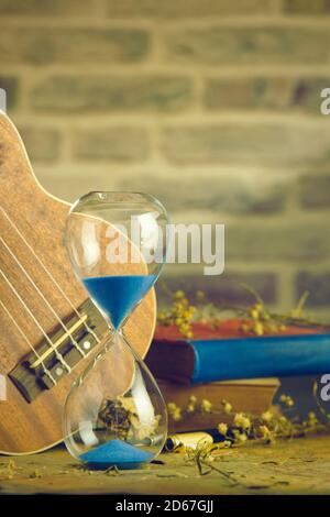A vintage hourglass and ukulele with an old book and brass pen on a wooden table and brick background in the morning. Closeup and copy space. The conc Stock Photo