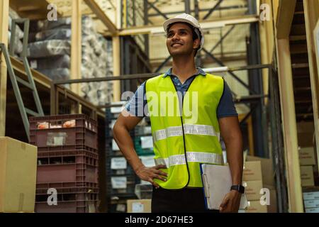 Young Indian industrial factory warehouse worker working in logistic industry indoor. Smiling happy man looking far away holding a clipboard checking item merchandise stock order in storehouse Stock Photo