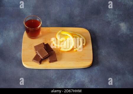 Light wooden board with a glass of cognac, chocolate pieces and lemon wedges on a gray background. Close-up. Stock Photo