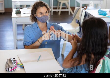 Female teacher and girl wearing face masks talking to each other through sign language in class Stock Photo