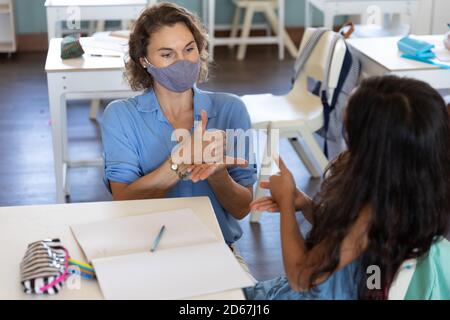 Female teacher and girl wearing face masks talking to each other through sign language in class Stock Photo