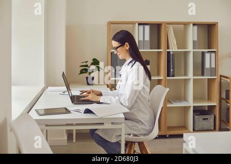 Smiling woman doctor sitting in medical clinic with laptop and typing something during online consultation Stock Photo
