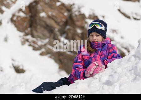 beautiful happy little girl in ski suit and wool hat with goggles lying on white snow in mountains during winter outdoor leisure christmas vacation Stock Photo