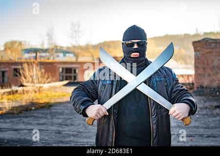 a bandit wearing black glasses and a mask stands with a crossed machete in his hands Stock Photo