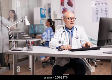 Elderly aged doctor using computer in hospital cabinet while young doctor talking with female patient on clinic corridor and nurse typing rapport on pc. Stock Photo