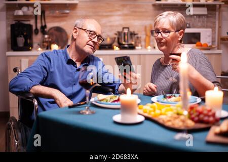 Man in wheelchair holding phone while having dinner with wife in kitchen. Scrolling and showing photos. Imobilized handicapped senior husband scolling on phone enjoying the festive meal. Stock Photo
