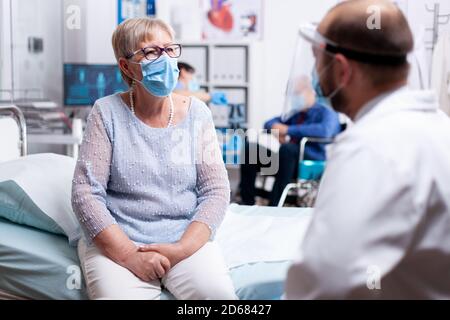 Elderly woman discussing with doctor during consultation in hospital examination room wearing face mask agasint covid-19. Global health crisis, medical system during pandemic, sick elderly patient. Stock Photo
