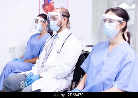 Medical personnel with face mask and visor against coronavirus sitting on chairs in hospital waiting area. Medic wearing stethoscope. Stock Photo