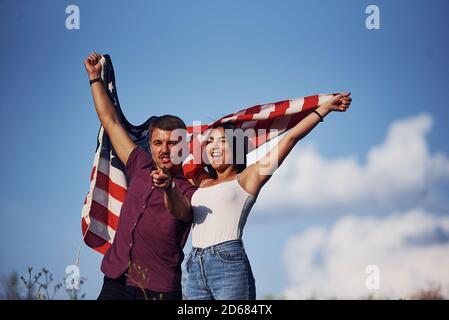 With hands up. Feels freedom. Beautiful couple with American Flag have a good time outdoors in the field Stock Photo