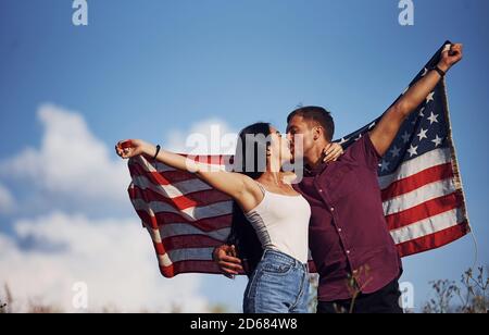 Kissing each other. Feels freedom. Beautiful couple with American Flag have a good time outdoors in the field Stock Photo