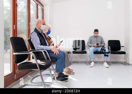 Senior couple with face masks sitting in a waiting room of a hospital together with a young man who is looking a a smart phone Stock Photo