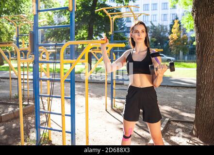 Muscular brunette pretty woman training arm using dumbbell in park, sports ground. Young female with elastic taping on body training outdoors. Rehabil Stock Photo