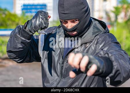 a black masked bandit with a knife in his hand stands in a fighting stance ready to attack and stares into the eyes of the victim Stock Photo