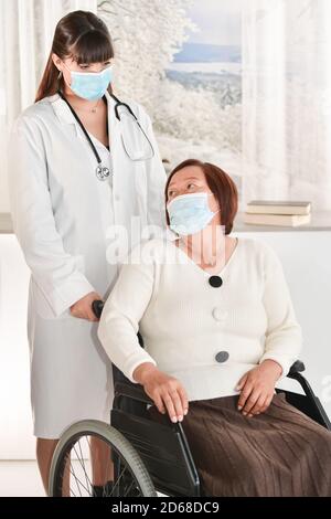 Mature asian woman in a wheelchair looking back at a young female doctor, both wearing surgical masks. healthcare and safety concept. Stock Photo
