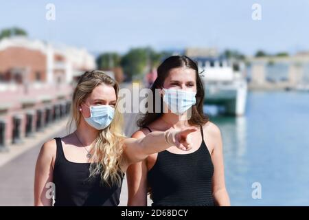 Two women wearing surgical masks pointing and looking on an out of focus background. Safety and leisure concept. Stock Photo