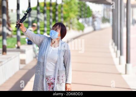 Mature asian woman using a face mask holding a small tripod with a compact camera outdoors on an out of focus background. Safety and leisure concept. Stock Photo
