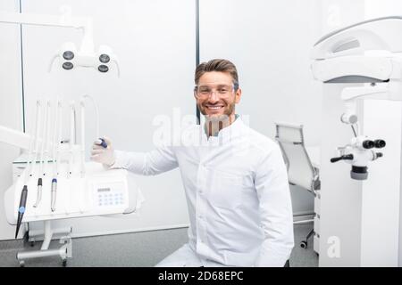 Confident male dentist sitting in a modern clinic near his dental chair. Dental hygienist wearing protective glasses smiling at the camera Stock Photo