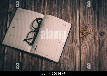 A pair of glasses folded on an open diary laid on a rustic wooden desk Stock Photo