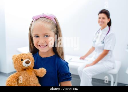 Smiling little girl with toy standing at the hospital ward. doctor is sitting on a background Stock Photo