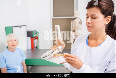 doctor Orthopedist points to the anatomical model of the foot. Arthritis, foot injuries and pain. On background senior patient Stock Photo