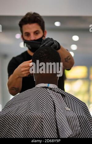 A young tattooed barber cuts a customers hair while wearing a face mask to conform to COVID-19 regulations Stock Photo