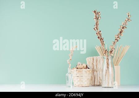 Contemporary interior in trendy mint menthe color with beige dry plant, flowers, bunch, basket and glass vases on soft light white wood table. Stock Photo