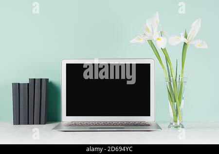 Fresh spring workplace for work and study with blank laptop display, black books and white iris bouquet in light green mint menthe interior on white w Stock Photo