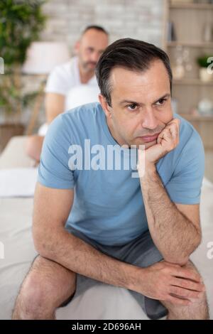 thoughtful man sitting on bed while boyfriend sleeping Stock Photo