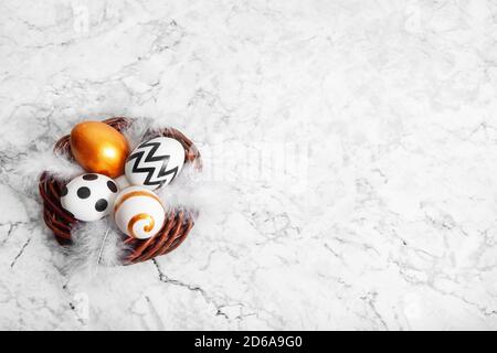 Happy Easter. Congratulatory easter background. Eggs painted gold and black in a nest with white feathers on marble background with copy space. Stock Photo